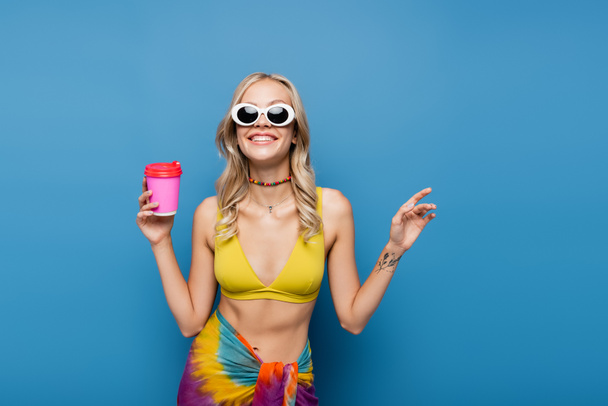 veselá mladá žena v slunečních brýlích a bikinách top drží papírový šálek izolované na modré  - Fotografie, Obrázek