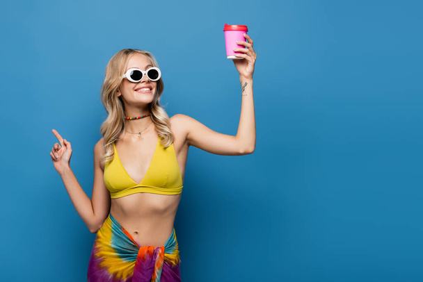 pleased young woman in sunglasses and bikini top holding paper cup on blue  - Foto, Bild