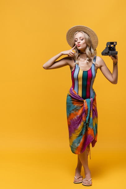 pleine longueur de femme blonde en chapeau de paille boudant lèvres et tenant caméra vintage sur jaune - Photo, image