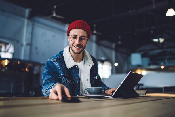 Positive handsome man in casual outfit surfing internet in smartphone while sitting at table with digital table and cup of drink and smiling in loft - Fotó, kép