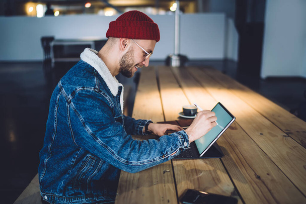 Seitenansicht eines jungen, gut aussehenden Mannes in lässigem Outfit, der mit einem modernen digitalen Gerät arbeitet und Stift benutzt, während er am Holztisch im Dachboden sitzt - Foto, Bild