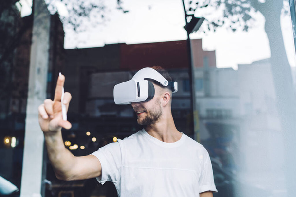 Cheerful young male wearing virtual reality glasses and enjoying innovative technologies while looking away and grinning brightly on dark blurred background - 写真・画像