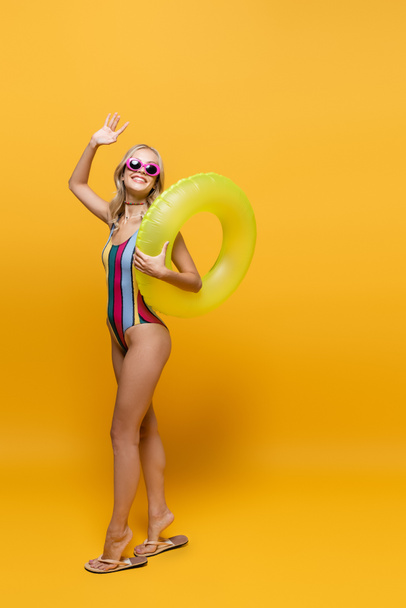 longitud completa de la mujer sonriente en gafas de sol y traje de baño que sostiene el anillo inflable y la mano ondulante en amarillo - Foto, imagen