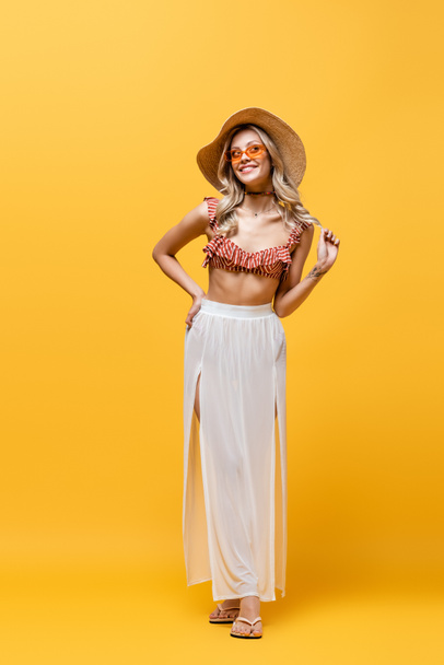 full length of smiling woman in ruffle bikini top and white skirt standing with hand on hip on yellow  - Foto, immagini