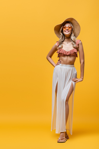 pełna długość uśmiechnięta kobieta w falbanki bikini top i biały spódnica pozowanie z ręki na biodrze na żółty  - Zdjęcie, obraz