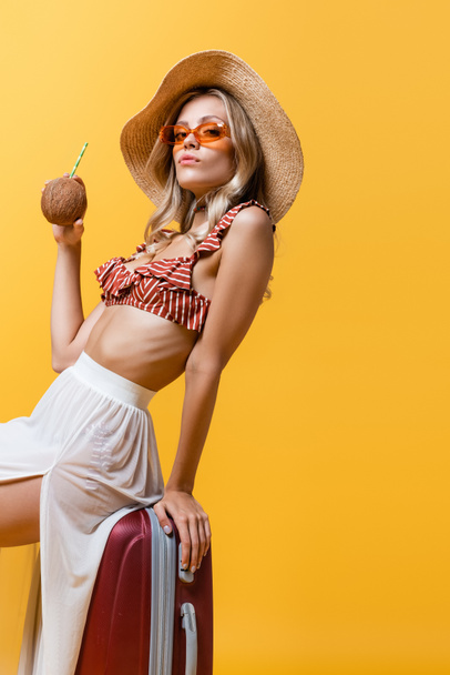 blonde woman in sun hat sitting on luggage and holding coconut with straw isolated on yellow  - Photo, Image