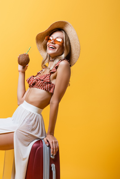 cheerful woman in sun hat sitting on luggage and holding coconut with straw isolated on yellow  - Photo, Image