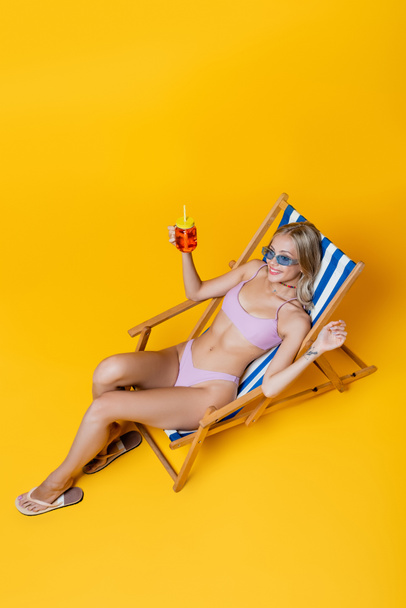 vue grand angle de jeune femme heureuse en maillot de bain assis sur la chaise longue avec cocktail sur jaune  - Photo, image