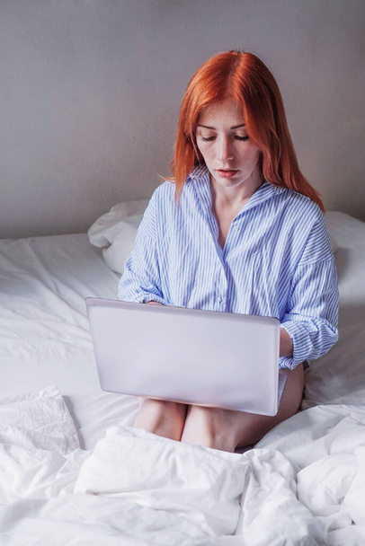 młoda kobieta siedzi w łóżku z laptopem na kolanach i wykonuje zdalną pracę z domu - Zdjęcie, obraz