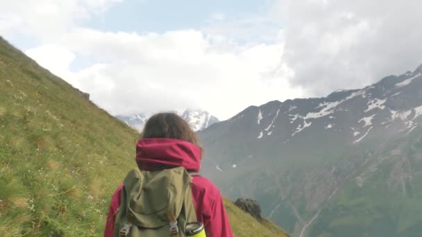 Een vrouwelijke toerist wandelt langs een groene helling op een bergpad tegen de achtergrond van besneeuwde toppen met een rugzak - Video