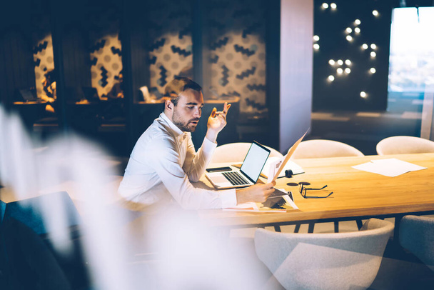 Blick aus dem Fenster eines ernstzunehmenden selbstbewussten Mannes in formeller Kleidung, der beim Start-up gestikuliert und Papierkram am Arbeitsplatz nachts überprüft - Foto, Bild