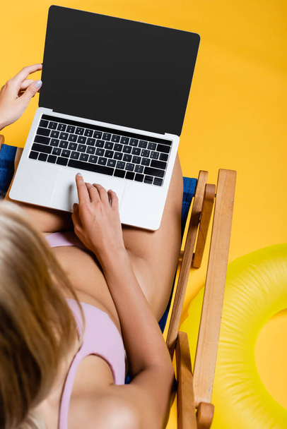 vista superior de la mujer rubia en traje de baño usando el ordenador portátil mientras está sentado en la silla de cubierta en amarillo  - Foto, imagen
