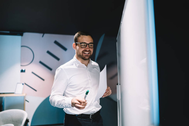 Intelligent smiling male in formal clothes and glasses with marker and paperwork going writing information on whiteboard for coworkers at office space  - Zdjęcie, obraz