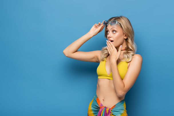 geschokt jonge vrouw in gele bikini top aanpassen zonnebril en weg te kijken op blauw - Foto, afbeelding