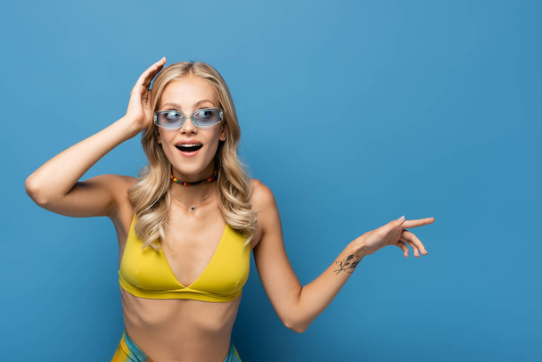 amazed young woman in bikini top pointing away isolated on blue - Photo, Image