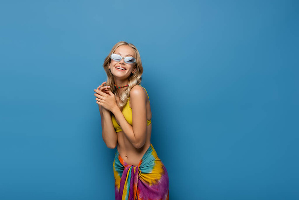 joyful young woman in sunglasses and beaded colorful necklace smiling isolated on blue - Photo, Image