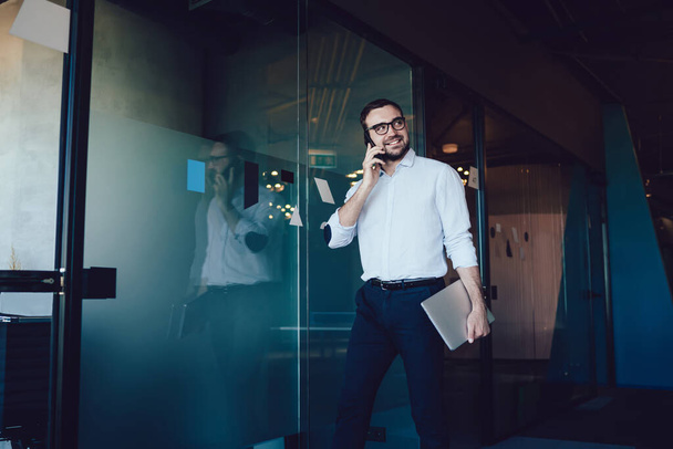 Content cheerful male in glasses in blue shirt and blue pants speaking on mobile phone in office opposite of glass door - Zdjęcie, obraz