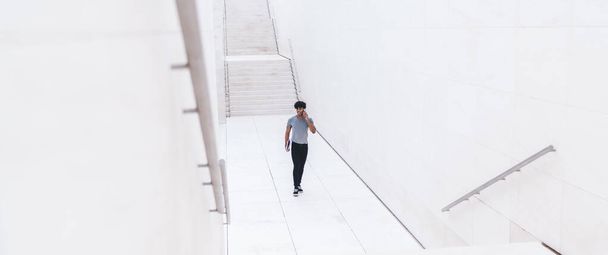 Casual modern male talking on smartphone while strolling in hallway of modern white tiled business center with stairs on street - Foto, Imagem