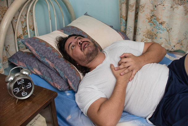 Ένας άντρας γύρω στα 30 υποφέρει είτε από καούρα είτε από καρδιακή προσβολή ενώ είναι ξαπλωμένος στο κρεβάτι.. - Φωτογραφία, εικόνα