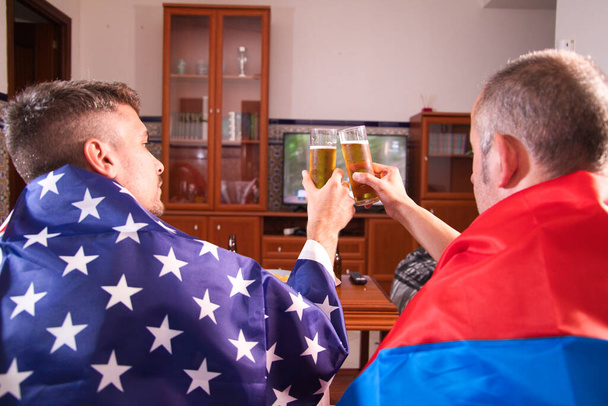 Two male friends and sports fans are watching a sporting event on television while toasting with a beer. Each of the men is carrying a flag on his shoulders. Sports Concept - Photo, Image