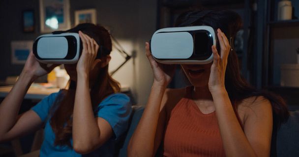 Ελκυστικές κυρίες της Ασίας απολαμβάνουν ευτυχισμένη στιγμή ψώνια σε απευθείας σύνδεση εμπειρία με γυαλιά εικονικής πραγματικότητας τοποθεσία ακουστικών στον καναπέ σαλόνι στο σπίτι σε σκοτεινή νύχτα. Χρήση με γυαλιά VR για ώρα ταινίας. - Φωτογραφία, εικόνα