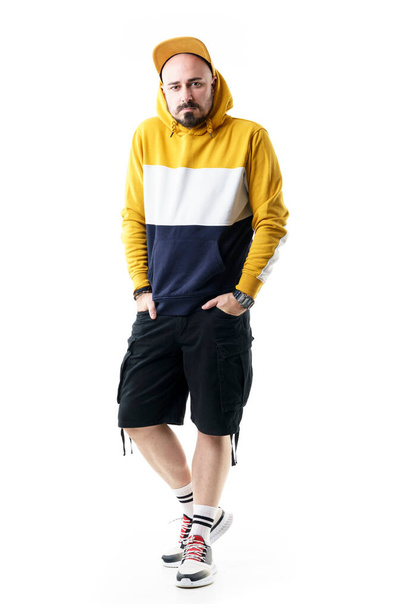 Young man wearing baseball cap, hooded sweatshirt and capri pants posing with hands in pockets. Full length portrait isolated on white background - 写真・画像