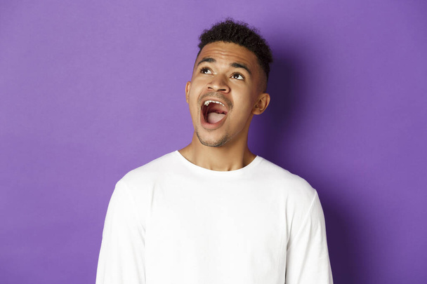 Крупный план изумленного африканско-американского молодого человека, открытого рта и говорящего вау, выглядящего очарованным в верхнем левом углу, стоящего на фиолетовом фоне с копировальным пространством - Фото, изображение