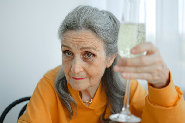 Bella vecchia nonna con i capelli grigi e il viso con le rughe seduti a tavola a casa sullo sfondo della finestra con un bicchiere di champagne, festa della mamma, felice pensionamento - Foto, immagini