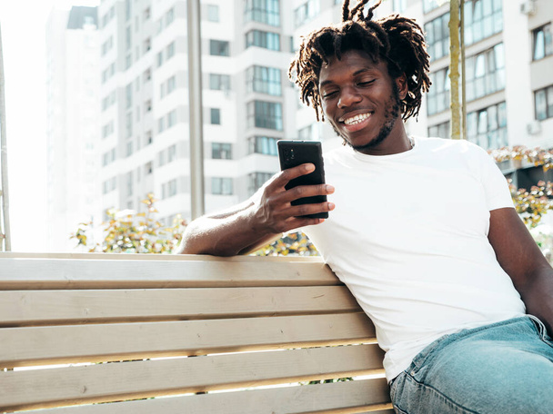 Handsome hipster model.Unshaven African man dressed in white summer t-shirt.Fashion male with dreadlocks hairstyle sitting at the bench in the street.Looking at smartphone screen, using cellphone apps - Foto, Imagem