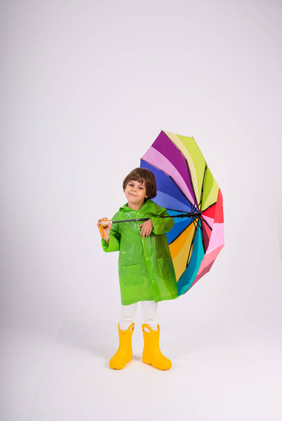 malý chlapec v zeleném plášti do deště a žluté gumové boty drží pestrobarevný deštník na bílém pozadí s kopií prostoru - Fotografie, Obrázek