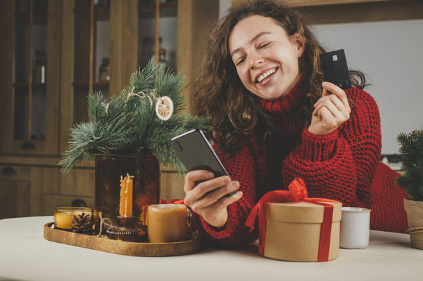 Красивая кудрявая молодая женщина, платящая онлайн банковской кредиткой за рождественские подарки, держа смартфон, глядя на экран. Женский клиент выбирает услугу, продукт в интернет-магазине с использованием 5g Интернет - Фото, изображение