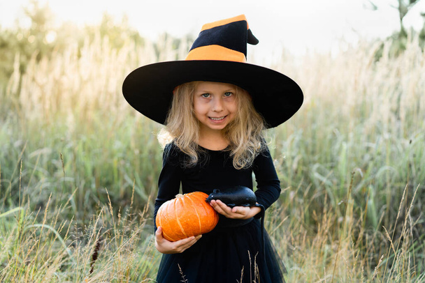 Egy szőke kislány fekete jelmezben és boszorkánykalapban, Halloween egy tökkel és egy patkánnyal a kezében, egy vidám gyerek..  - Fotó, kép