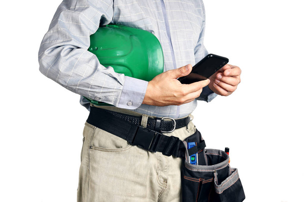 Builder με εργαλεία και σκληρό καπέλο κατέχει smartphone στα χέρια του, απομονώνονται σε λευκό φόντο - Φωτογραφία, εικόνα