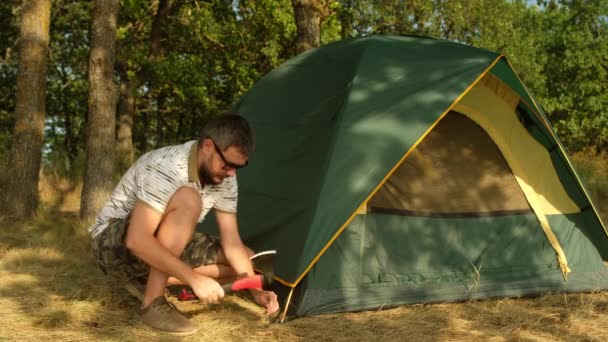 Un uomo con la barba allestisce una tenda nel campo turistico. Martelli femminili peg con un'ascia. - Filmati, video