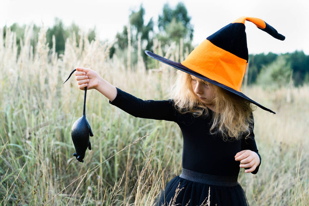 little blonde girl in a black suit and a witch hat with a black rat in her hands, Halloween, a fun holiday. - Photo, Image