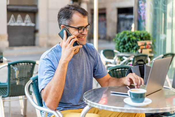Mladý latinský muž pracující spokojeně s mobilním telefonem a počítačem venku. - Fotografie, Obrázek