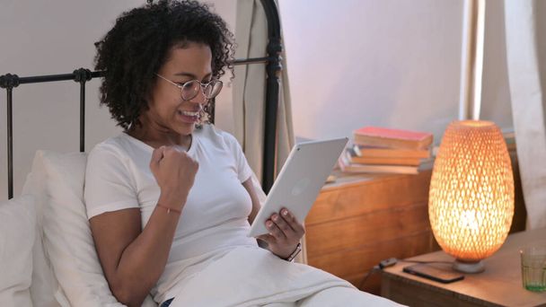 jong Afrikaans vrouw vieren succes op tablet in bed - Foto, afbeelding