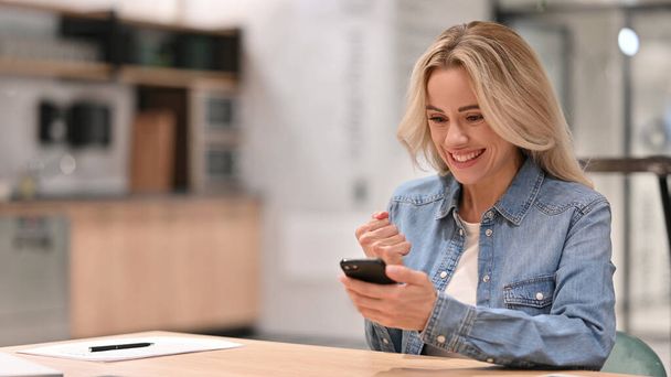 Junge Gelegenheitsfrau, die bei der Arbeit mit dem Smartphone jubelt, Erfolg - Foto, Bild
