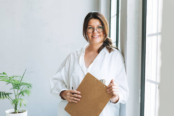 Portrait of Young smiling brunette woman plus size near window with pen and documents in hands in bright modern office - Photo, Image