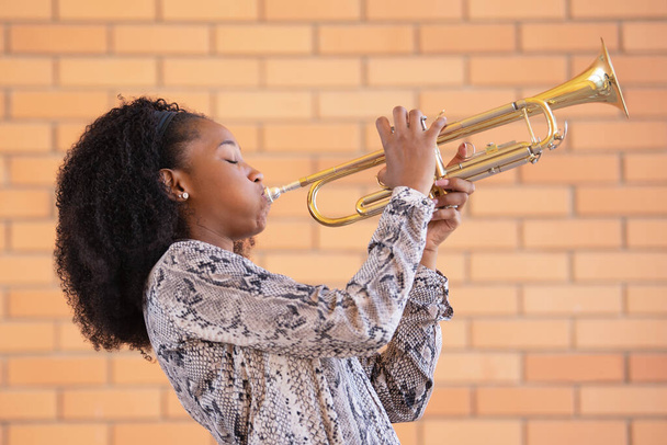 jong afro amerikaans vrouw spelen de trompet met haar ogen dicht op een baksteen muur achtergrond - Foto, afbeelding