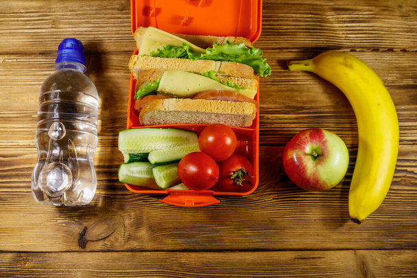 Eine Flasche Wasser, Apfel, Banane und Lunchpaket mit Sandwiches und frischem Gemüse auf einem Holztisch. Ansicht von oben - Foto, Bild