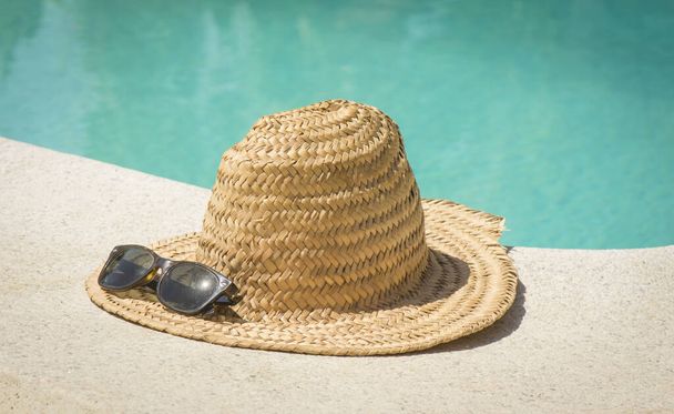 A vintage straw hat with sunglasses at the border of a swimming pool in summer - Foto, afbeelding