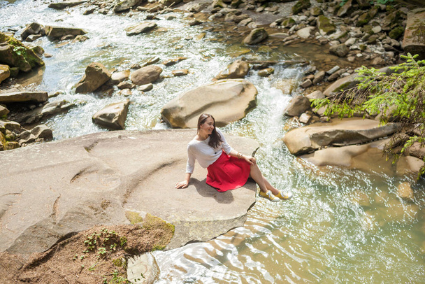 Girl in a blouse and skirt sitting near a mountain river - Φωτογραφία, εικόνα