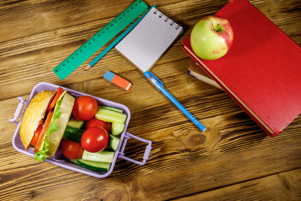 Okul konseptine geri dönelim. Okul gereçleri, kitaplar, elma ve beslenme çantası içinde hamburgerler ve ahşap bir masada taze sebzeler. Üst görünüm - Fotoğraf, Görsel