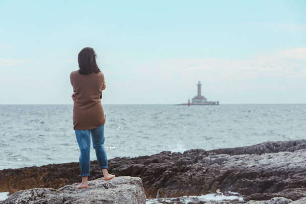 женщина прогуливается по скалистому морскому пляжу в мокрых джинсах маяка на заднем плане. ветреная погода. летние каникулы. Беззаботная концепция
 - Фото, изображение