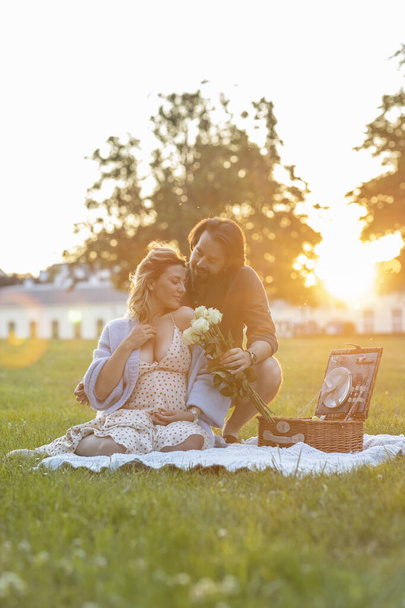 Beautiful pregnant young couple having a picnic at sunset in the grass - 写真・画像