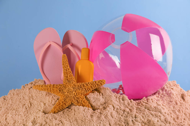 Beach ball, sunscreen, flip flops and starfish on sand against light blue background - Фото, изображение