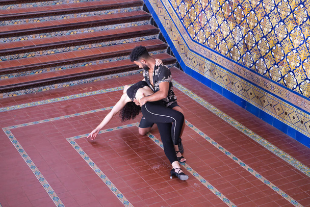 Multi-ethnic young couple making figures while dancing bachata sensually outdoors next to a beautiful tiled wall. - Фото, изображение