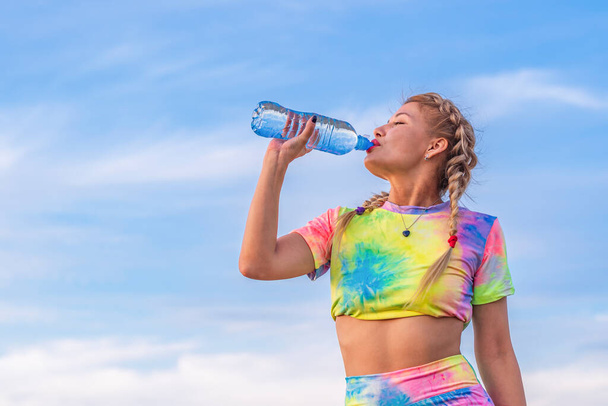 Young woman in a bright multicolored tracksuit drinks water from a transparent plastic bottle against a blue summer sky. Outdoor sports. - Photo, Image