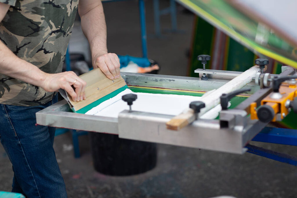 Διαδικασία εκτύπωσης μεταξοτυπίας Serigraphy στο εργοστάσιο ρούχων. Χρώματα κορνίζας, squeegee και πλαστισόλης - Φωτογραφία, εικόνα
